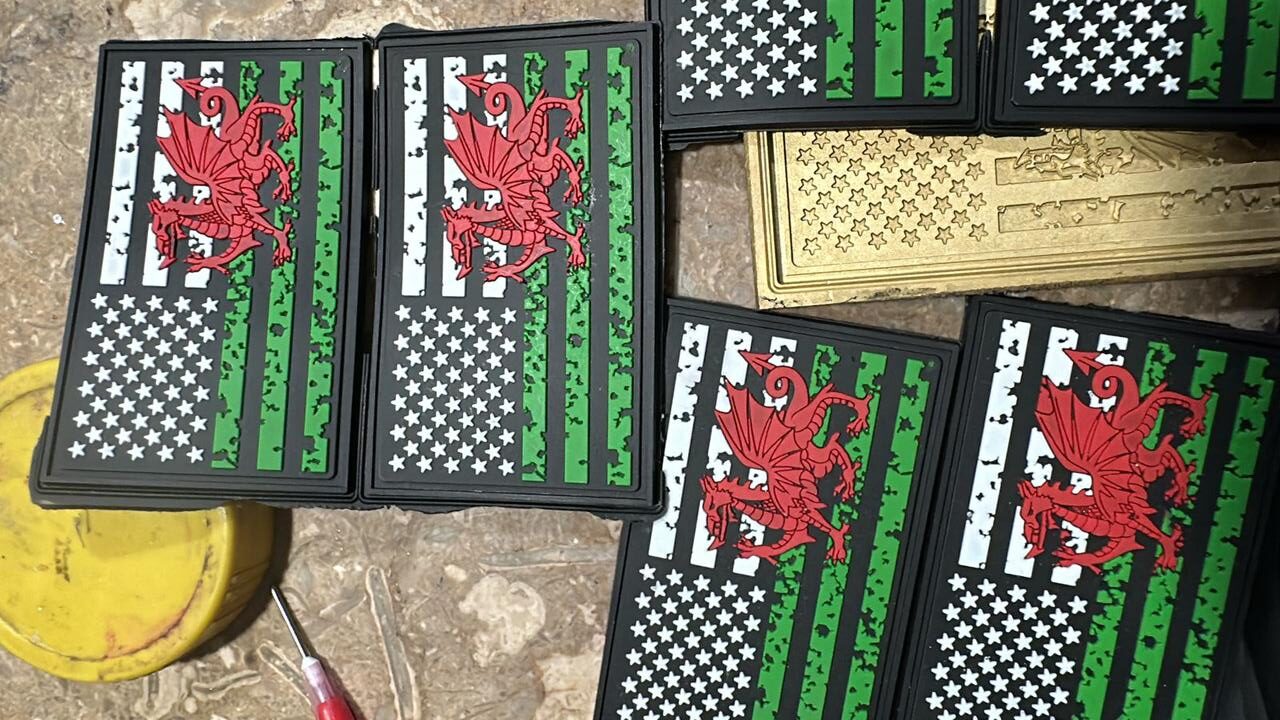 Stacked patches featuring a stylized American flag in black, white, and green with a red Welsh dragon overlay in the center